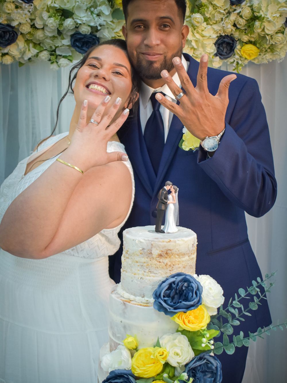 Novios mostrando sus anillos frente al pastel y un fondo de flores azules y amarillas