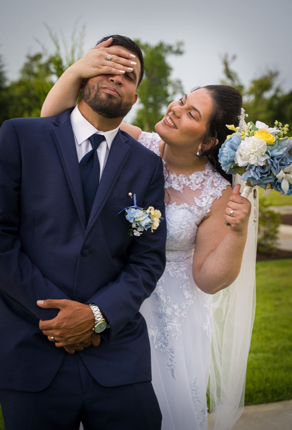 Novia tapándole los ojos al novio con una sonrisa, ramo azul y amarillo